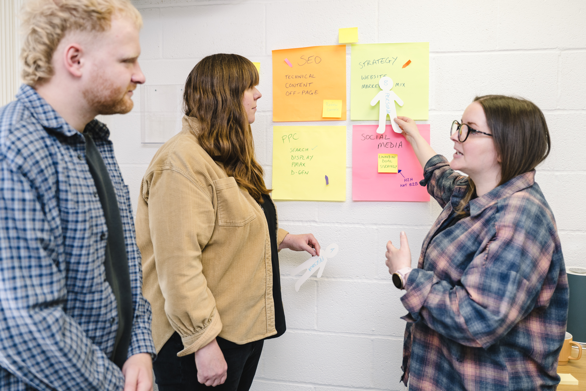 three office workers dressed in casual clothes stood up in the office, pointing at a customer persona surrounded by marketing strategies on post it notes