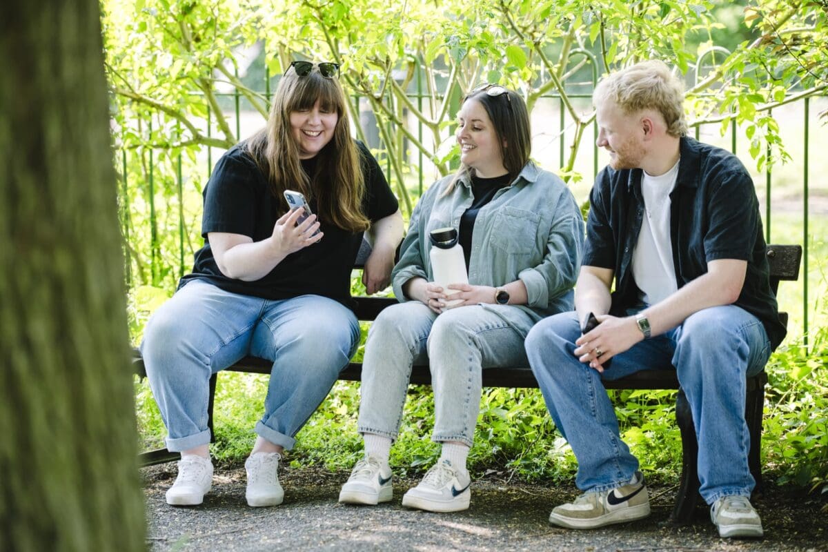 three office workers looking at a phone and smiling while sitting on a bench outside in the sun,