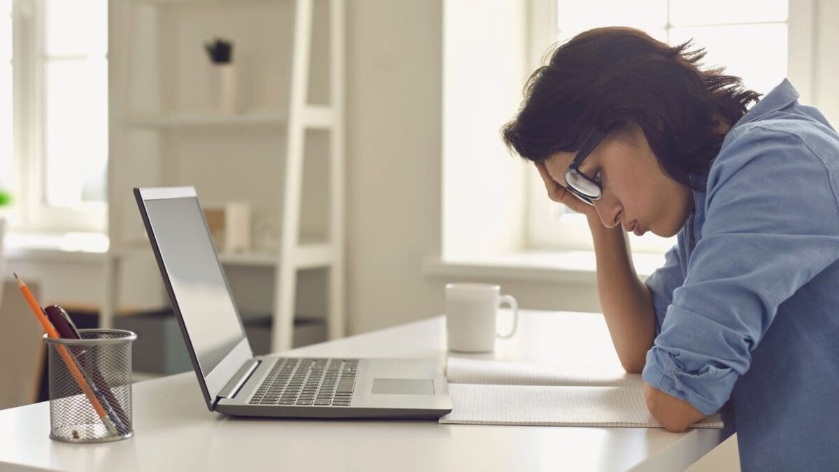 A tired woman wearing glasses sits at a desk with her head resting on her hand, looking down at a notebook beside an open laptop.