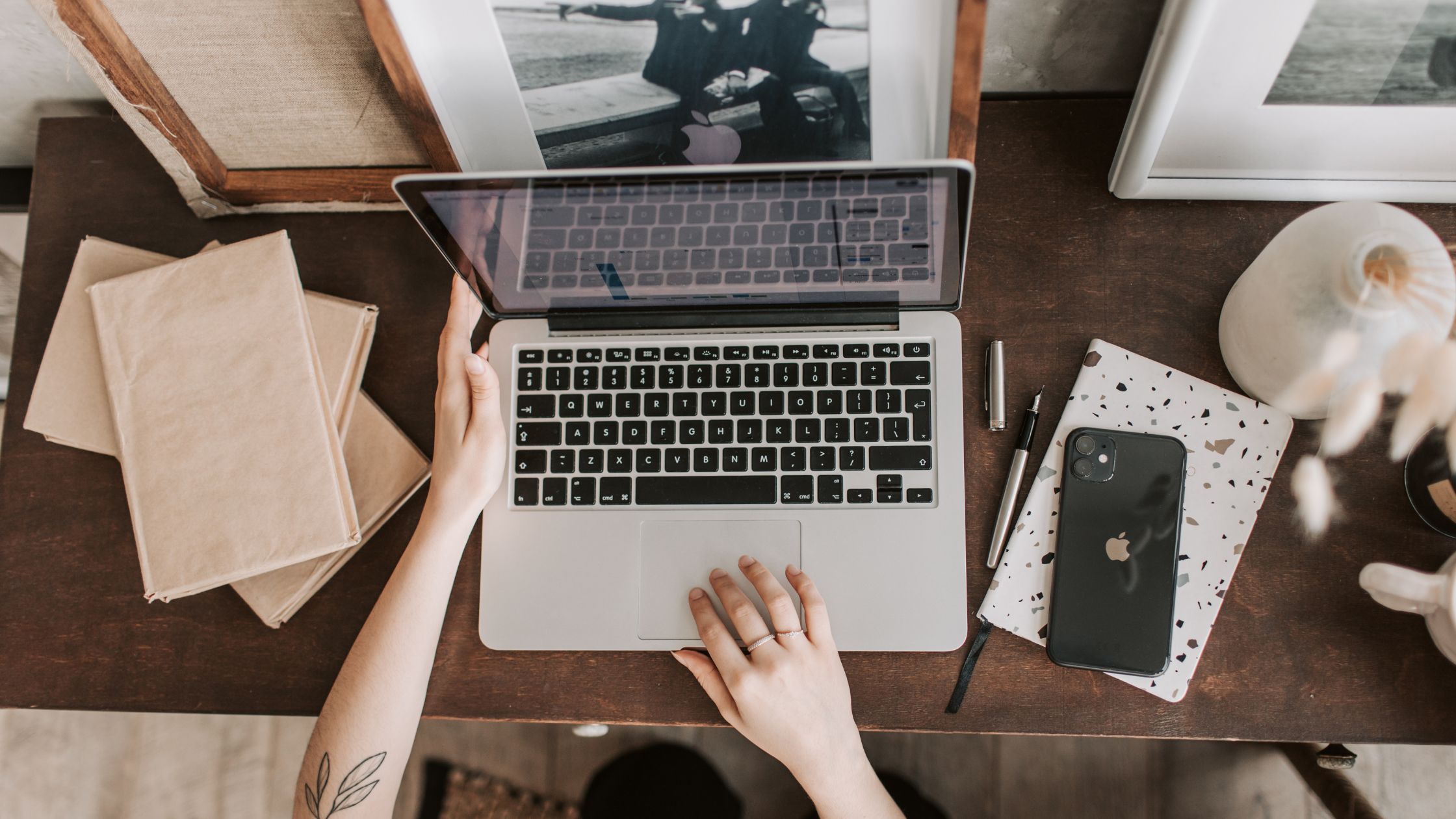 A person uses an open laptop at a wooden desk, surrounded by stacked books, a notebook, a smartphone, a pen, and a small vase.