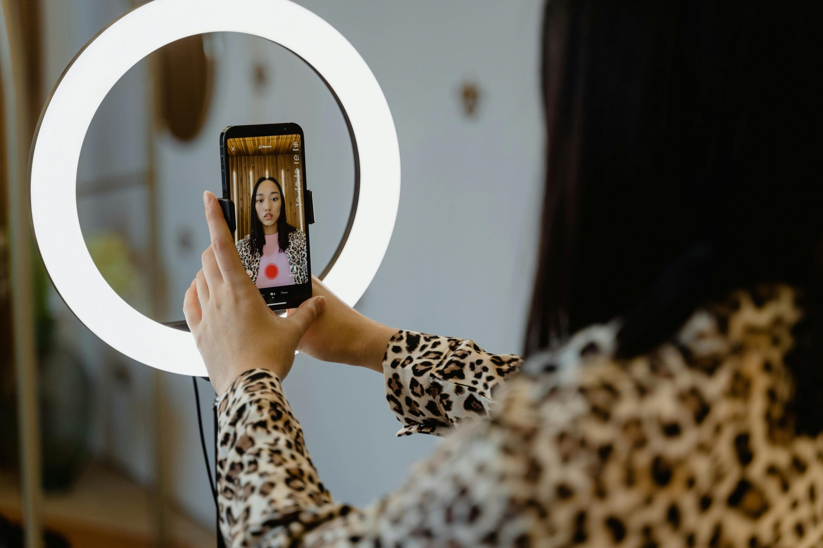A woman records a video of herself on a smartphone positioned inside a bright ring light.