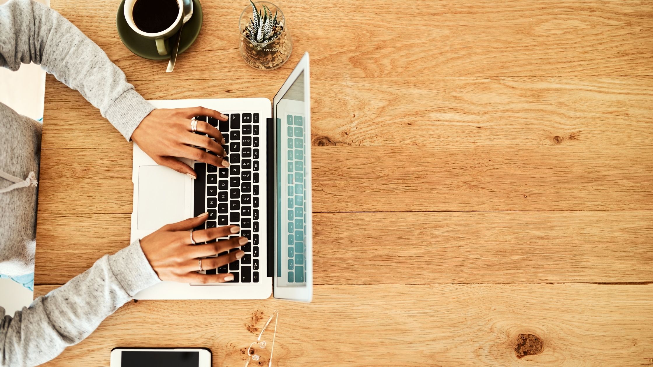 Overhead view of a person typing on a laptop at a wooden desk, with a coffee cup, smartphone, and small plant nearby.